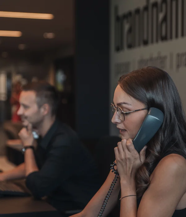 Seminarraum in Südtirol zum Mieten gesucht? Der Brandnamic Campus ist ideal Frau spricht am Telefon in einem modernen Büro mit einem Mann am Computer im Hintergrund