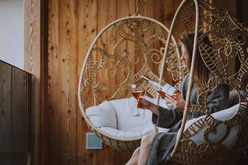 Wein, Hotel und Marketing – ein leckeres Trio Frau sitzt im Hängesessel mit Weinglas und Flasche in Holzzimmer.