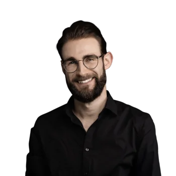 Julian Mair | Senior Software Developer Junger Mann mit Bart und Brille, schwarzes Hemd, lächelnd vor weißem Hintergrund