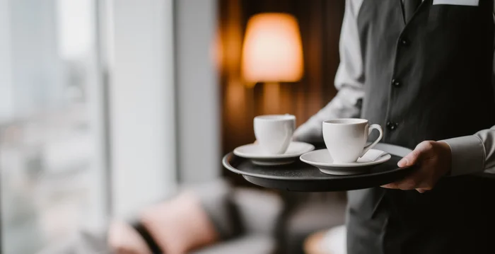Marketing für Hotels – eure Hotelmarketing-Agentur aus Südtirol Kellner trägt zwei Tassen Kaffee auf Tablett in gemütlichem Hotelzimmer