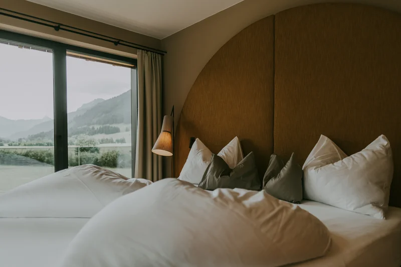 Wein, Hotel und Marketing – ein leckeres Trio Gemütliches Bett mit vielen Kissen und Bergblick durch Fenster