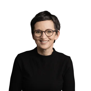 Helga Karbon | Campus Support Frau mit kurzen dunklen Haaren, runder Brille und schwarzem Pullover lächelnd