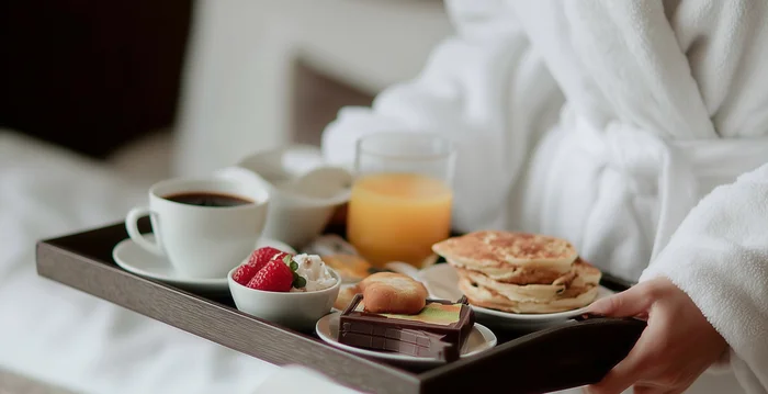 Marketing per hotel – la vostra agenzia di marketing per hotel in Alto Adige Persona in accappatoio con vassoio colazione con caffè, succo, pancake e frutta