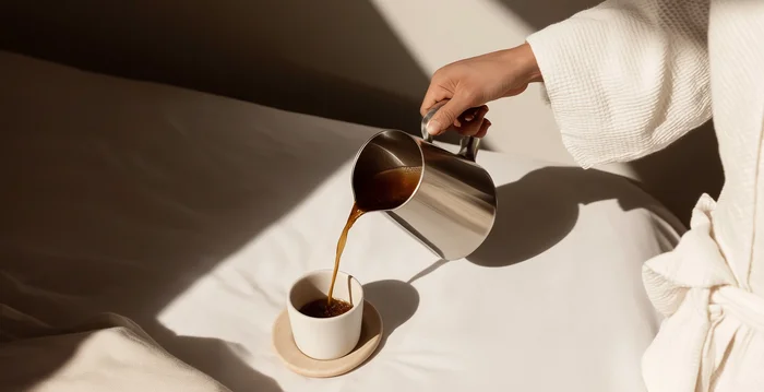 Hotelberatung: Experten für die Hospitalitybranche Hand gießt Kaffee aus Metallkanne in Tasse auf Bett mit Tageslicht