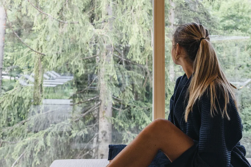 Erfolgreiches Marketing aus Brixen Frau in Bademantel schaut auf grüne Bäume durch großes Fenster